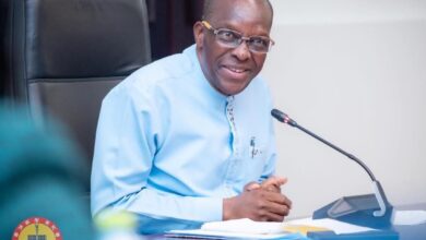 Speaker of parliament speaking into a microphone while seated at a meeting table, smiling during a formal discussion.