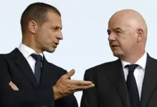 Aleksander Čeferin (left), wearing a dark suit and a dotted tie, gestures with his hand while speaking to Gianni Infantino (right), who is bald and wearing a matching dark suit.