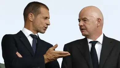 Aleksander Čeferin (left), wearing a dark suit and a dotted tie, gestures with his hand while speaking to Gianni Infantino (right), who is bald and wearing a matching dark suit.