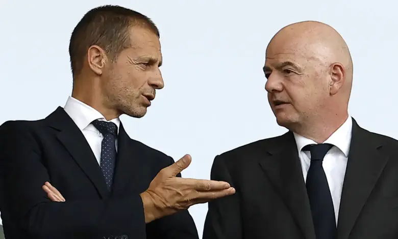 Aleksander Čeferin (left), wearing a dark suit and a dotted tie, gestures with his hand while speaking to Gianni Infantino (right), who is bald and wearing a matching dark suit.